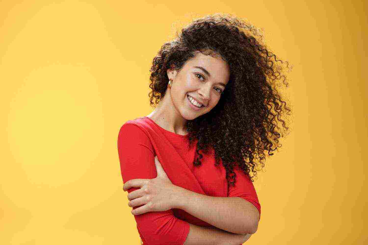 waist up shot tender feminine gentle woman with curly hairstyle combed right side tilting head smiling flirty making romantic gazed camera hugging herself yellow background