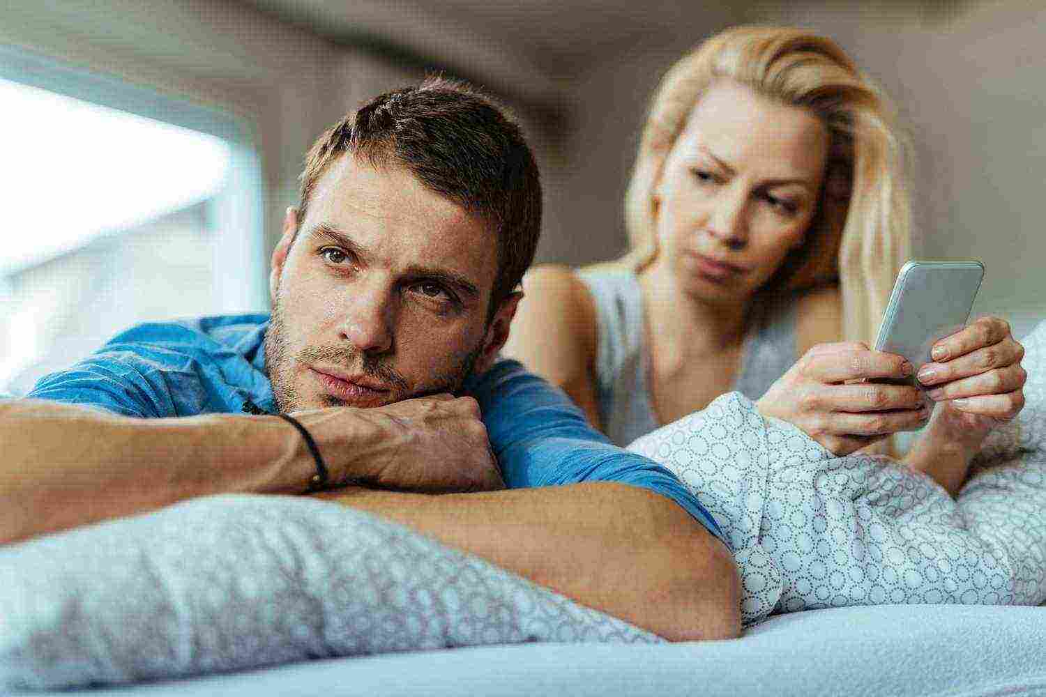 displeased man lying bed ignoring his girlfriend who is text messaging mobile phone
