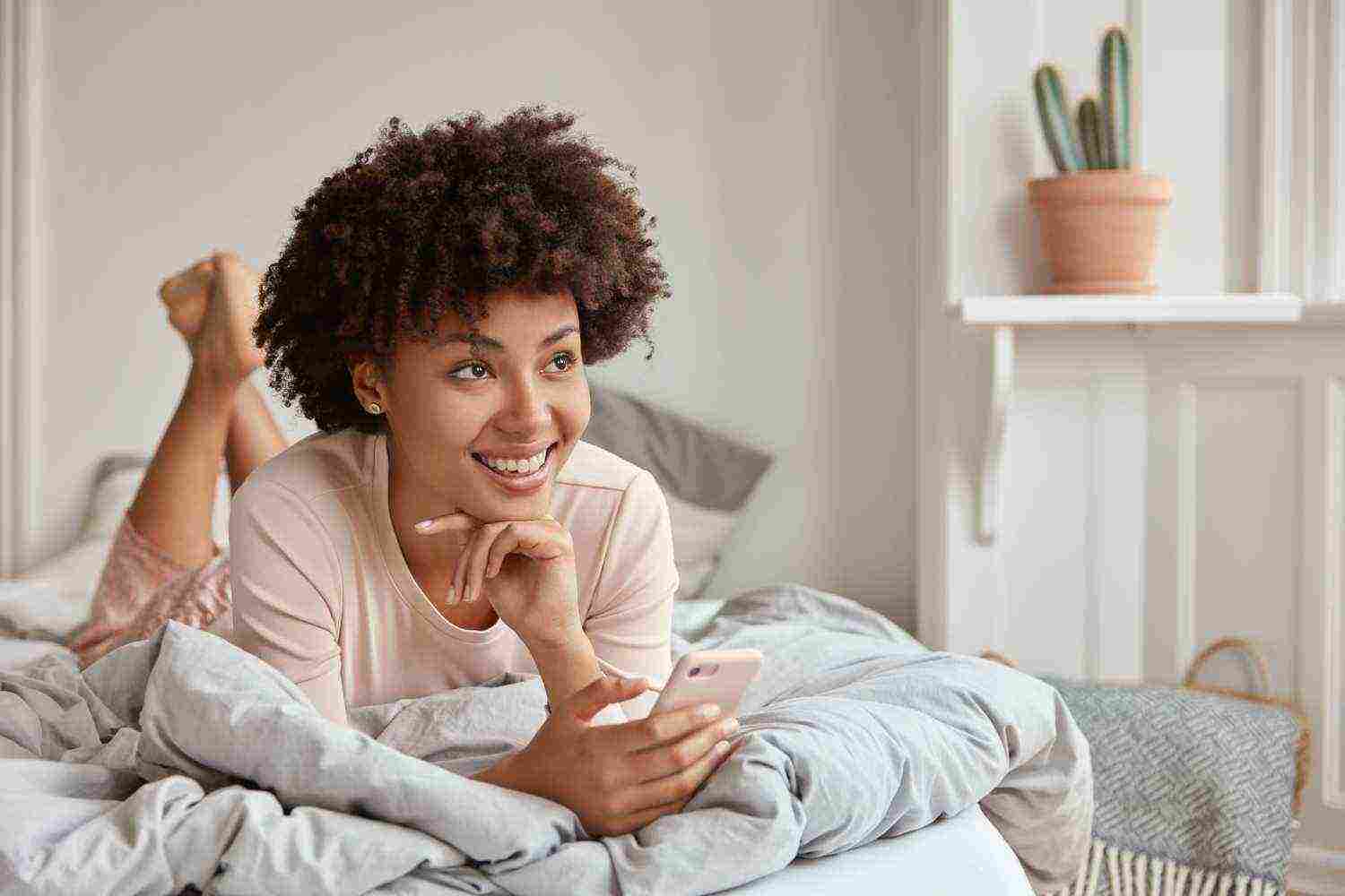 relaxed dark skinned female with afro hairstyle keeps legs raised dressed pyjamas holds modern cellular has dreamy expression browses internet sends text messages enjoys calm atmosphere