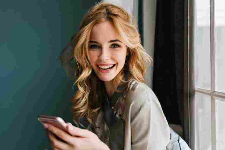 young cheerful woman with long wavy hair with pink smartphone hands looking smiling sitting window wearing silk pajama turquoise wall curtains