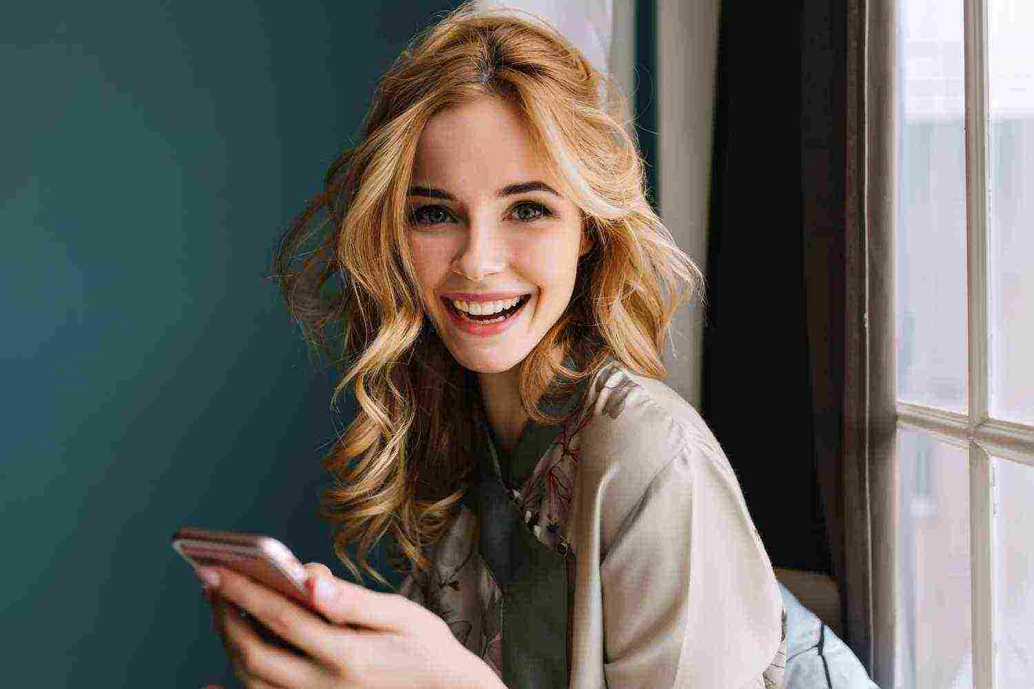 young cheerful woman with long wavy hair with pink smartphone hands looking smiling sitting window wearing silk pajama turquoise wall curtains