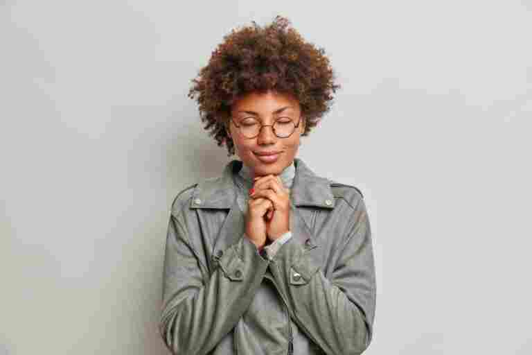 calm hopeful young woman with curly hair keeps hands together prays hopes better enjoys peaceful atmosphere wears spectacles grey jacket