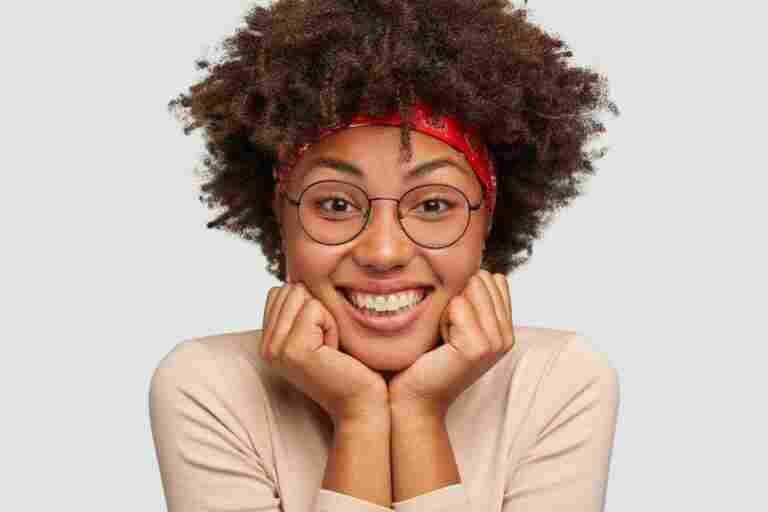 close up portrait happy black lady holds chin with both hands glad that everything is okay wears round glasses has curly hair listens funny story from interlocutor positive emotions concept