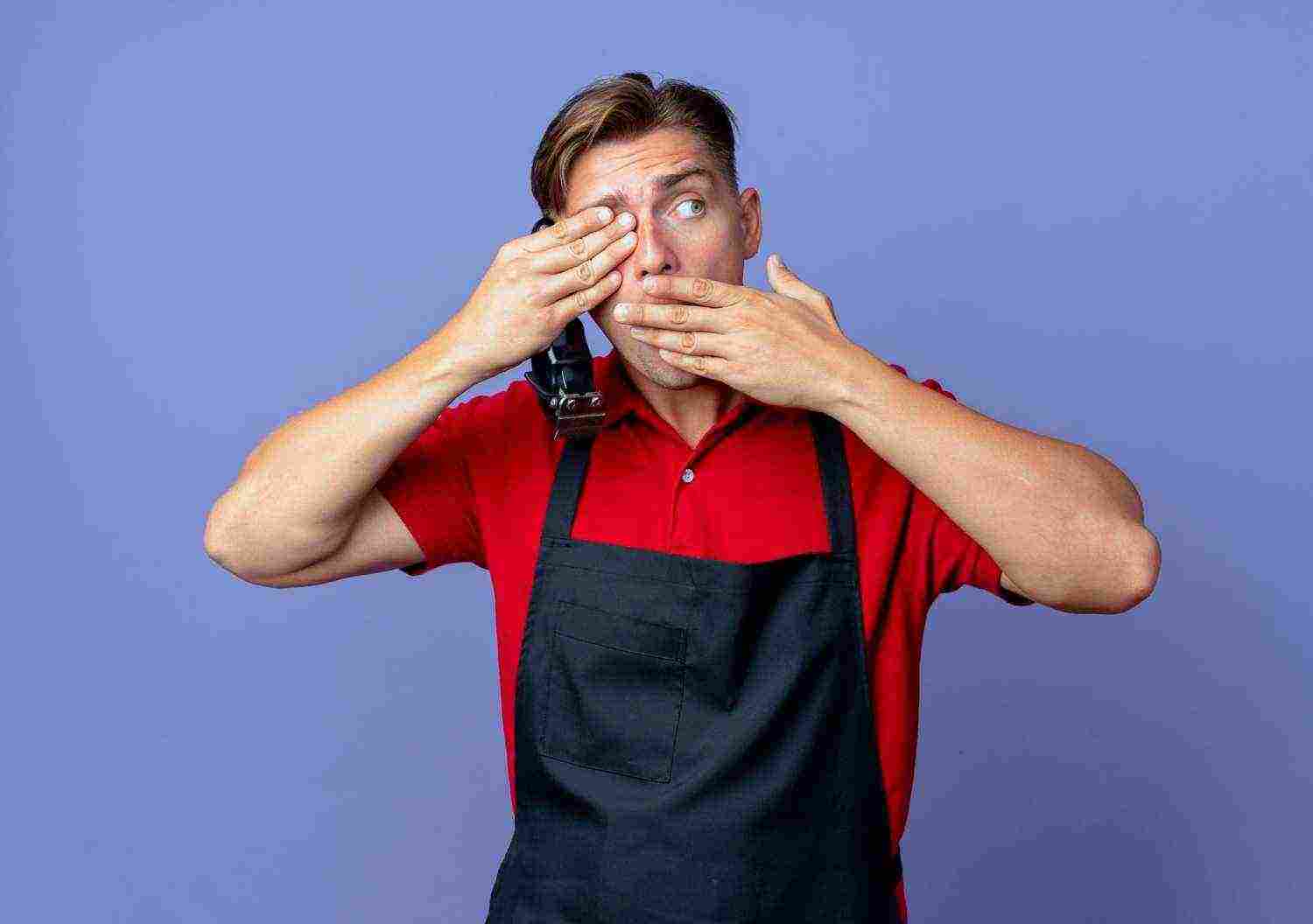 young shocked blonde male barber uniform closes eye with hand puts hand mouth holding hair clipper looking side isolated violet space with copy space