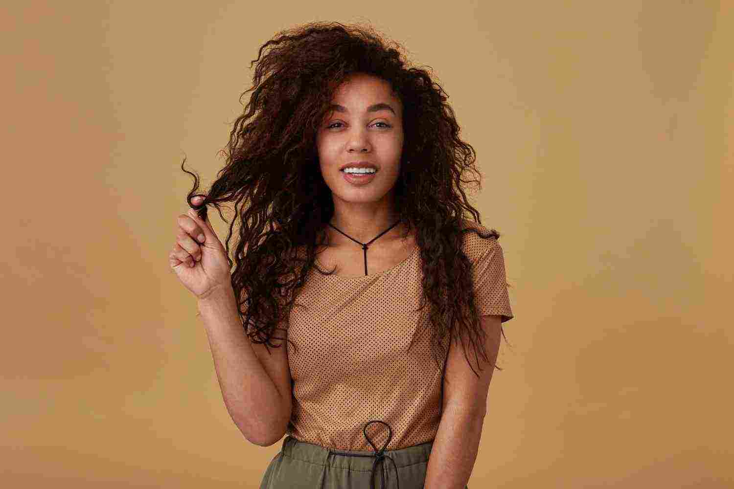 cheerful young pretty dark skinned lady twisting her brown curly hair raised finger smiling gladly while posing beige casual wear