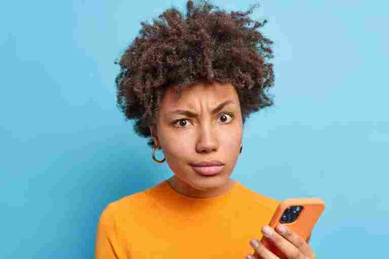 discontent afro american woman smirks face looks with dissatisfied expression holds modern cellular reads negative news online wears orange jumper isolated blue wall technology concet