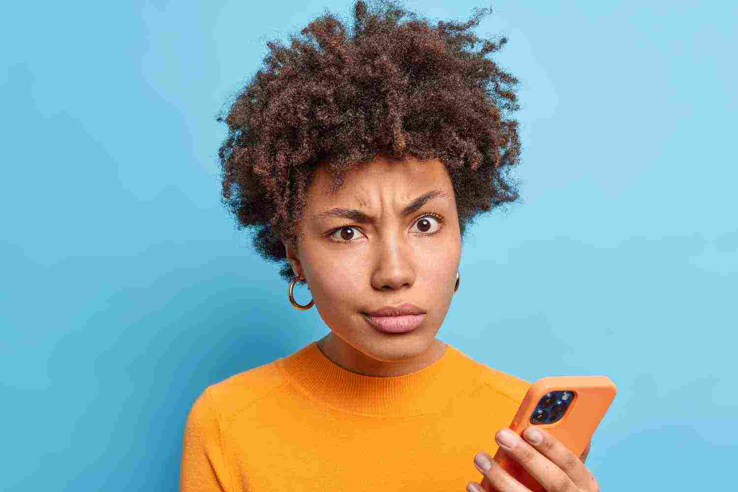 discontent afro american woman smirks face looks with dissatisfied expression holds modern cellular reads negative news online wears orange jumper isolated blue wall technology concet