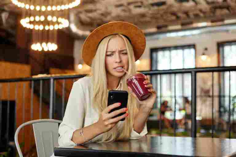 displeased blue eyed young long haired blonde lady keeping mobile phone hand looking screen with pout frowning face showing her white perfect teeth