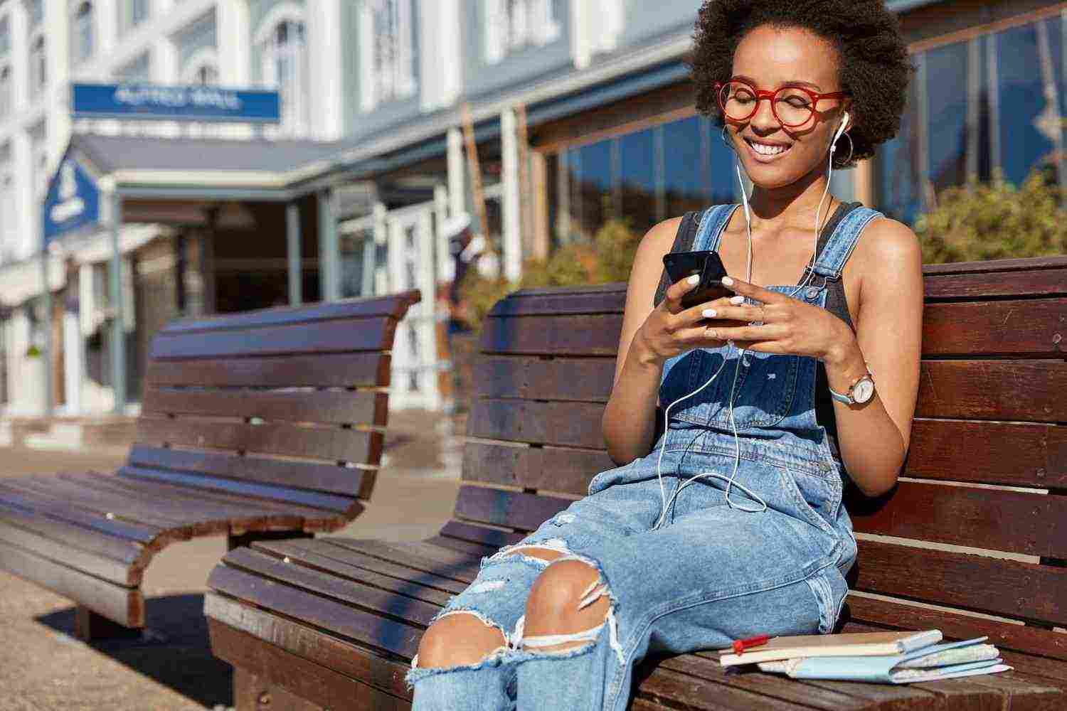 photo cheerful black woman holds cellular types text messages uses earphones listens music dressed ragged overalls models outdoor enjoys playlist modern technologies online communication