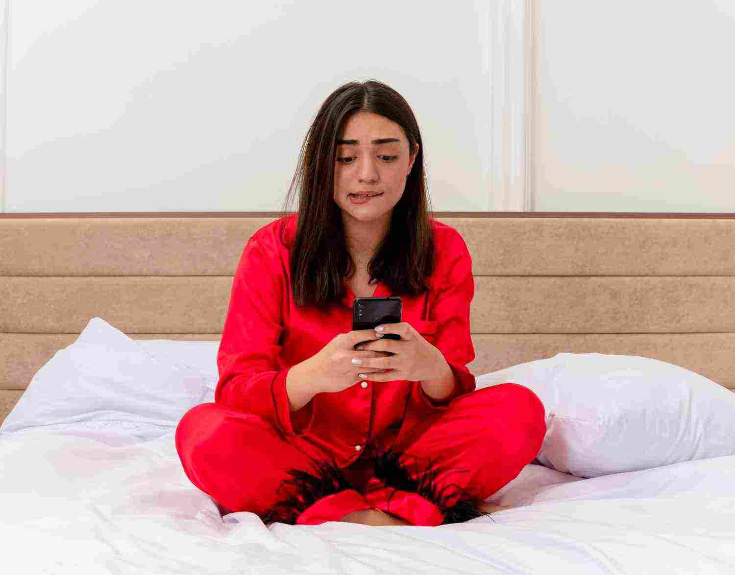 young beautiful woman red pajamas sitting bed using smartphone looking confused nervous bedroom interior light background