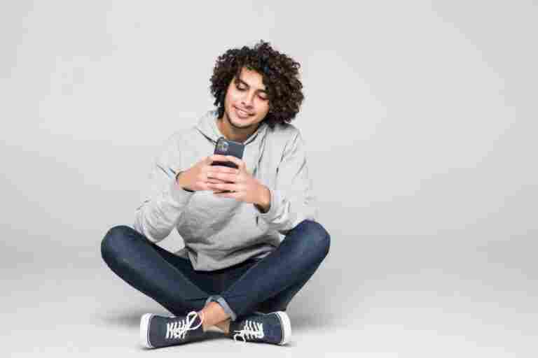 young curly man sitting floor sending message isolated white wall