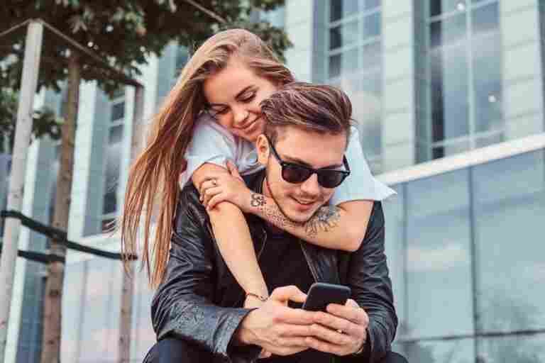 young fashionable couple relaxing together near skyscraper pretty girl cuddling her boyfriend while he using smartphone