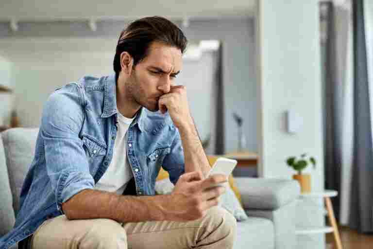 young worried man sitting sofa reading text message mobile phone