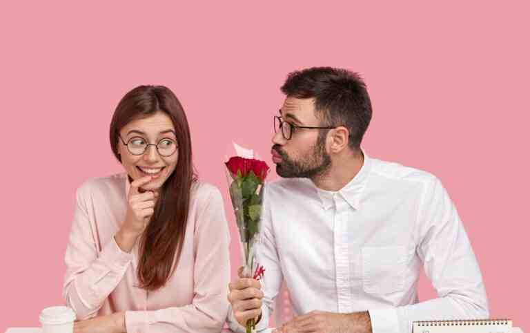 male boss fell love with young pretty colleague gives beautiful red roses folds lips making kiss happy lady receives compliment flowers sits desktop office against pink wall