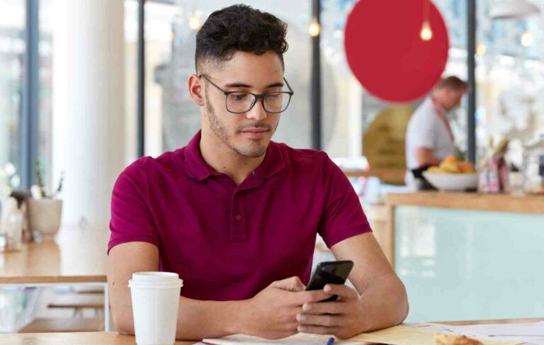 photo handsome man has satisfied expression holds modern cell phone sits cozy cafeteria watches video connected wireless internet drinks takeaway coffee people technology concept e1691315511692