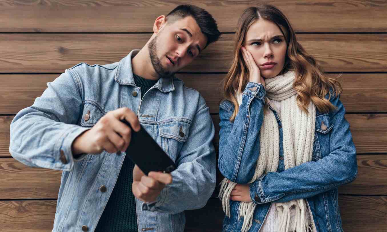 upset charming girl with long scarf looking away while posing wooden wall carefree caucasian man using his smartphone