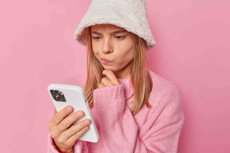 woman looks smartphone screen reads notification receives message from unknown person wears white hat jumper isolated pink has doubtful expression