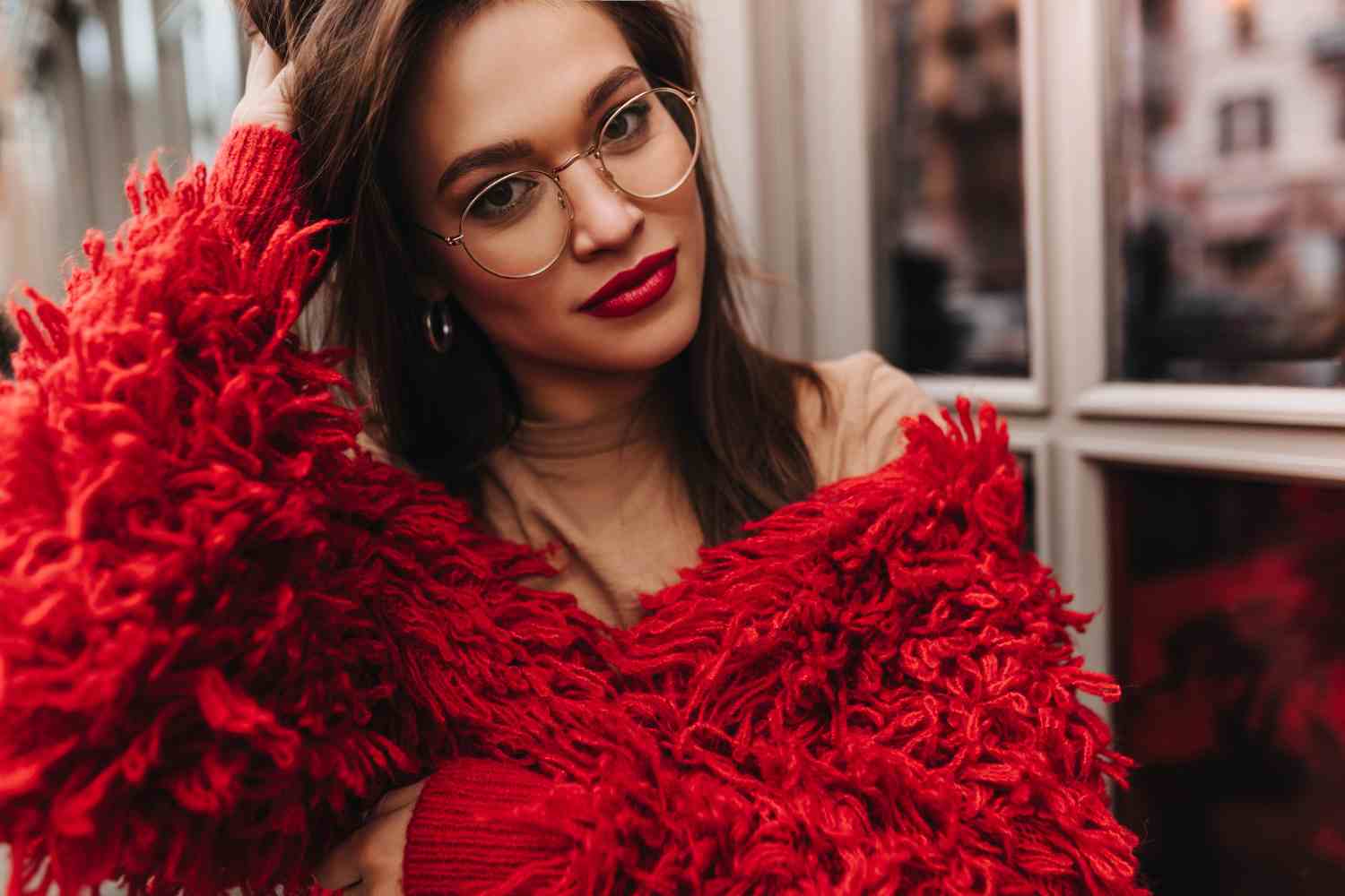 attractive brown eyed brunette woman with red lipstick dressed bright knitted jacket beige top looking into camera through glasses against windows
