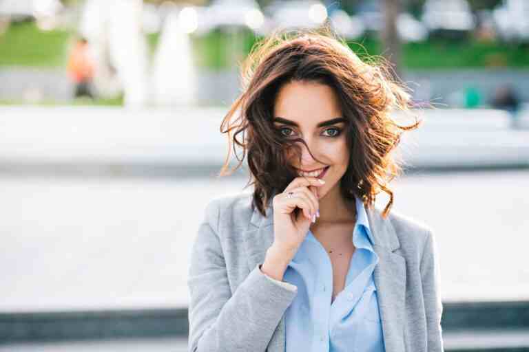 closeup portrait cute brunette girl with short hair walking city she wears blue shirt gray jacket she keeps hand lips smiling camera