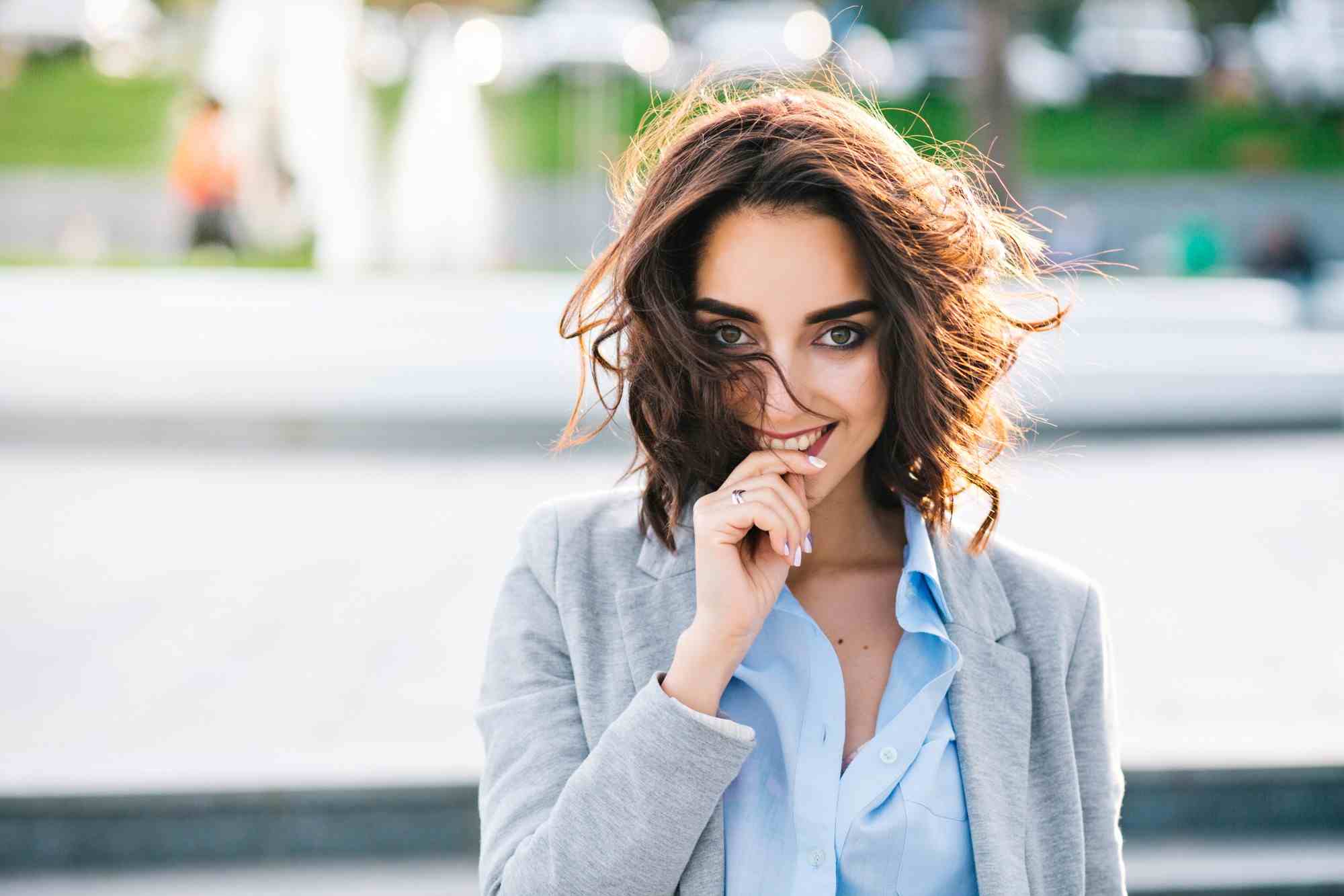 closeup portrait cute brunette girl with short hair walking city she wears blue shirt gray jacket she keeps hand lips smiling camera