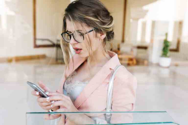 portrait young pretty woman stylish business lady surprised looking smartphone holding silver phone hands standing hall she has white short manicure wearing stylish pink jacket