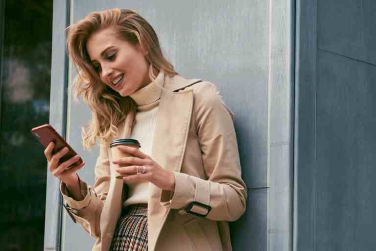 pretty smiling blond girl stylish trench coat with coffee go joyfully using cellphone outdoor