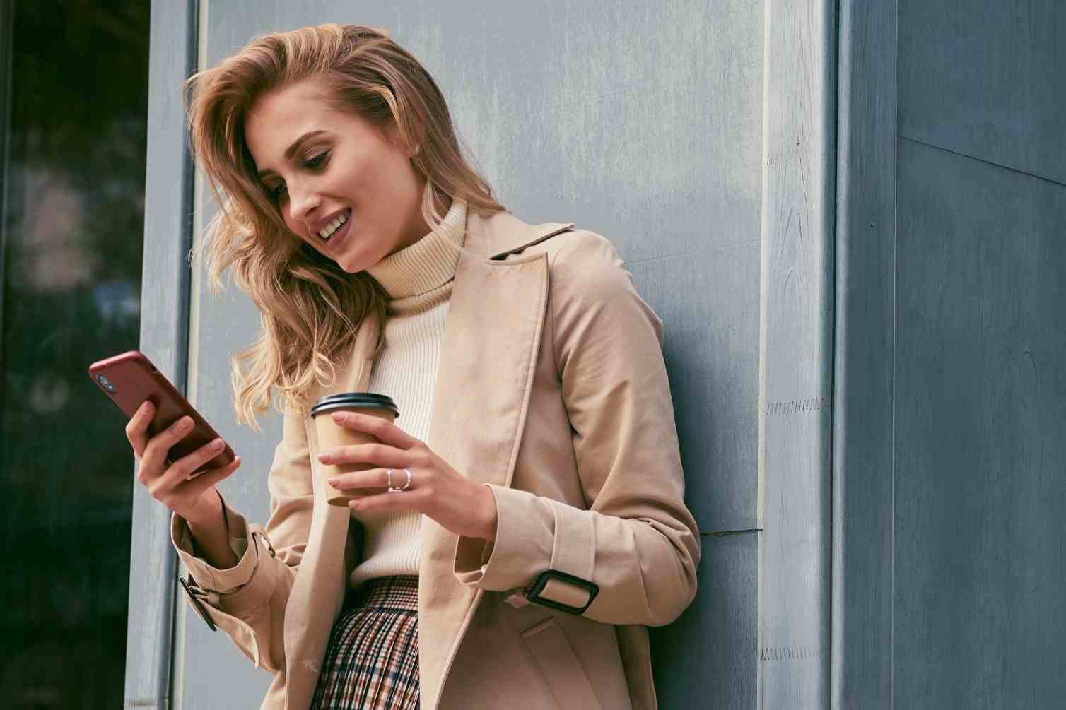 pretty smiling blond girl stylish trench coat with coffee go joyfully using cellphone outdoor