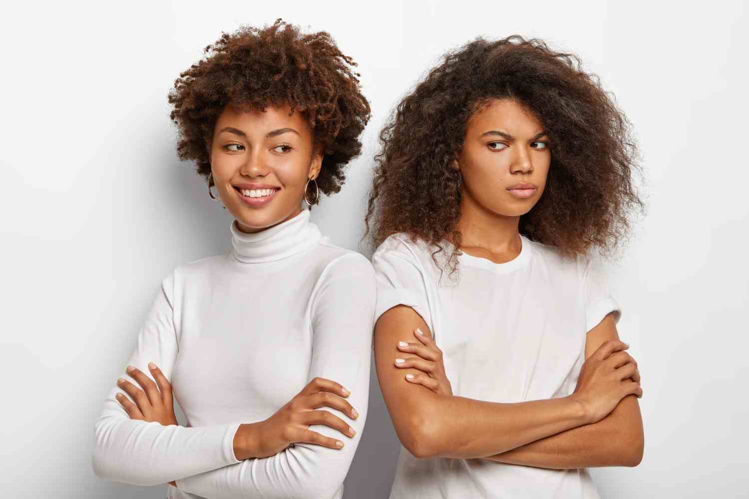 smiling dark skinned woman keeps hands crossed stand shoulder shoulder with best friend who has gloomy upset expression wear white clothes express different emotions